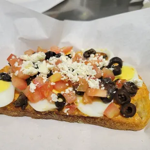 a piece of bread with eggs, tomatoes, olives, and cheese