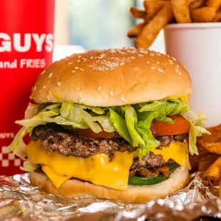 A close-up photograph of a Five Guys cheeseburger, soft drink in red Five Guys cup, and regular order of fries sitting on a t...