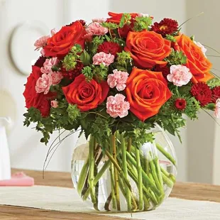 A vibrant bouquet of orange roses, red carnations, pink mini carnations, and lush greenery beautifully arranged in a round gl...