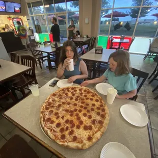 two girls sitting at a table with a large pepperoni pizza