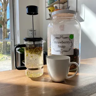 A French press infuses tea in a sunbeam. There is a white mug beside it and a jar of tea leaves labeled &quot;strawberry snap.&quot;