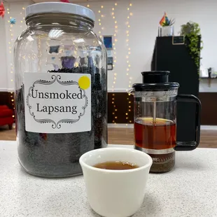 A jar of tea leaves labeled &quot;Unsmoked Lapsang&quot; sits on a counter beside a white teacup and a French press full of dark brewed tea.