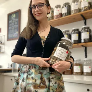 A brunette woman in a dinosaur dress hold a jar of tea leaves labeled &quot;Happy Place&quot; and smiles.