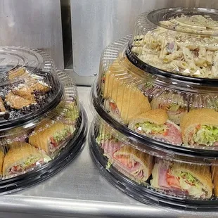 A counter showcases three trays filled with assorted sandwiches from Genova's To Go catering, perfect for any gathering.