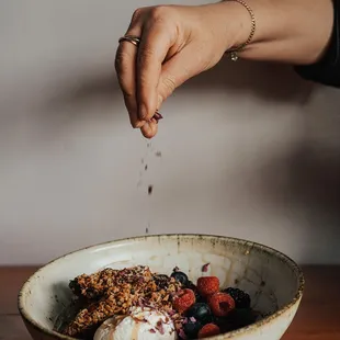 a hand sprinkling granola over a bowl of fruit