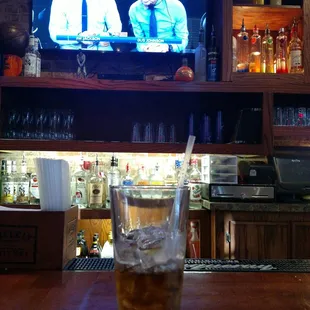 a glass of iced tea on a bar with a television in the background
