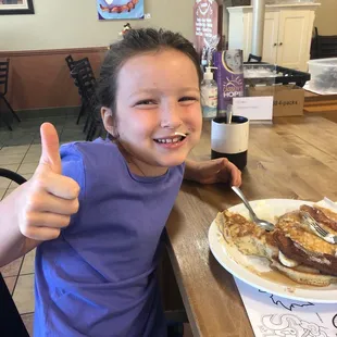 a young girl sitting at a table with a plate of pancakes