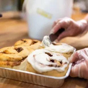 a person spreading icing onto a tray of cinnamon rolls
