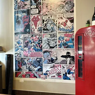 a coca cola machine in front of a wall covered with comics