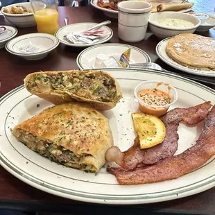 Sausage breakfast burrito with a side of bacon. In the background, amazing home fries, creamy grits, and excellent pancakes.