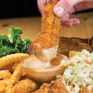 a hand dipping a piece of chicken tenders into a bowl of