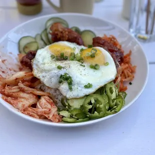 Korean fried chicken on rice and topped with an egg, sliced pickles, chopped carrots, kimchi, and sliced jalapeño peppers.
