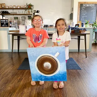 two young girls holding a painting of a cup of coffee
