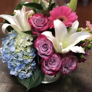 Fresh garden cluster of lavender roses, hot pink gerbera daises, blue hydrangeas, and white lilies in a glass vase.
