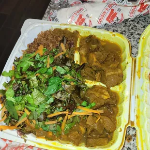 Goat with salad on top of rice in a To Go container.