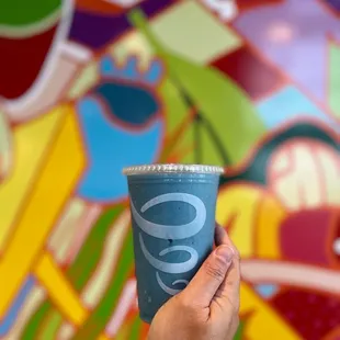 a hand holding a blue cup in front of a colorful mural