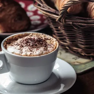a cup of cappuccino with a basket of croissants in the background