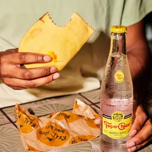 a person holding a piece of bread and a bottle of soda