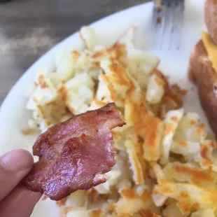 a hand holding a piece of bacon over a plate of potatoes