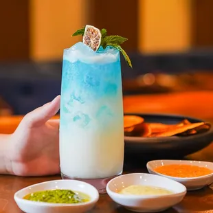 a hand holding a blue drink with a mint leaf garnish