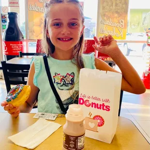 a little girl sitting at a table with a bag of donuts