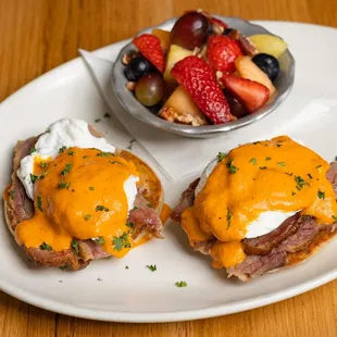 two eggs benedicts on a plate with a bowl of fruit