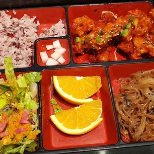 Clockwise from to left: purple rice, popcorn chicken, japchae, orange slices, soy ginger salad, and pickled radish at the center.