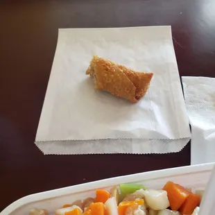 Egg roll served on a styrofoam plate and a to-go bag.