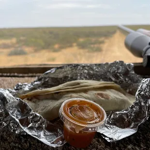 a burrito wrapped in tin foil with a jar of sauce