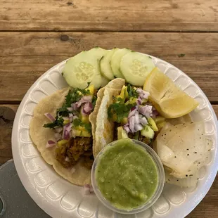 Al Pastor tacos topped with pineapple, red onions, and cilantro. With a side of cucumber, lemon, grilled onion, and green guacamole salsa.