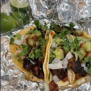 Three tacos al pastor with cilantro, onion, limes, and various sauces.