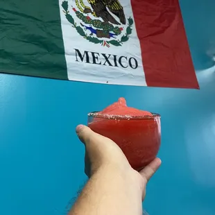 a hand holding a drink in front of a mexican flag