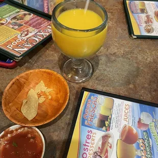 a glass of orange juice, a bowl of salsa, a bowl of chips and a plate of chips