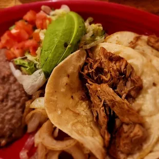 Three carnitas tacos with grilled onions beans lettuce tomato and avocado slices