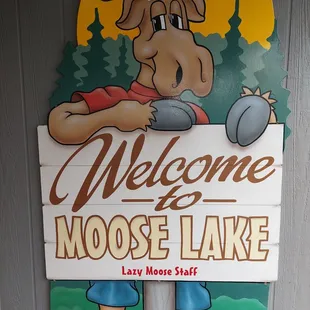 a moose holding a sign that says welcome to moose lake