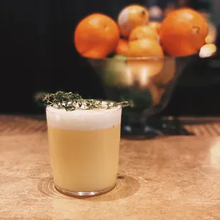 a drink in a glass on a table with oranges in the background