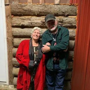 a man and woman standing in front of a log cabin