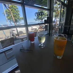 Huckleberry cider, vanilla bean cider, seven seas hazy IPA, and a Jones soda on a table with Alki beach and waterfront in the background.