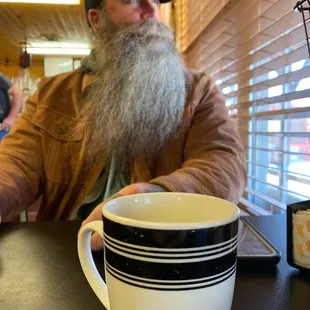 a bearded man sitting at a table with a cup of coffee