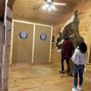 a man and a woman standing in a room with darts on the wall