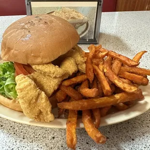 Fried catfish sandwich with lettuce, tomato and tartar sauce on a toasted bun with a side of sweet potato fries.