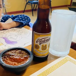 a little girl sitting at a table with a bowl of chili and a bottle of beer