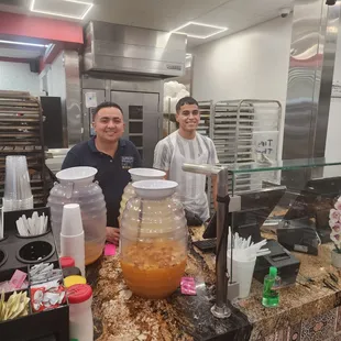 two men standing behind a counter with a jar of liquid