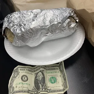 a hot dog wrapped in aluminum foil and sitting on a plate next to a hundred dollar bill