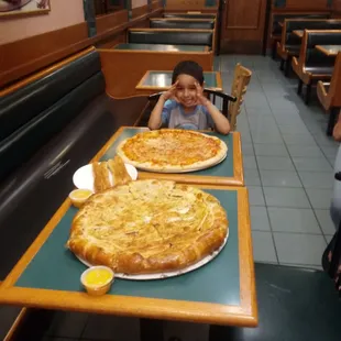 a boy sitting at a table with two large pizzas