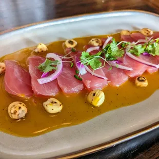 Tuna Tiradito. $16.99. Sashimi tuna with aji amarillo pepper, onions, cilantro, roasted corn nuts, and fresh citrus. Nikkei cuisine.