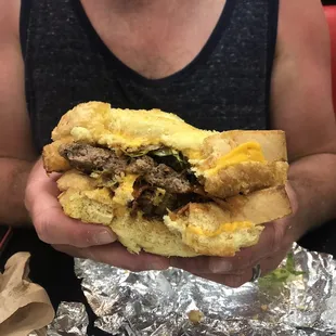 Goliath burger 2 beef patties, cheese, lettuce tomatoes, sautéed onions, mad sauce, between two grilled cheese sandwiches.