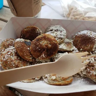 a person holding a wooden spoon in a box of powdered sugared donuts