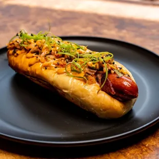 The Glizzy! All beef hotdog, wrapped in bacon &amp; topped with kimchi relish, chili crisp aioli, green onions. All inside a milk bread bun.