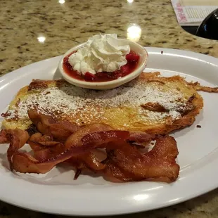 Egg Nog croissant French Toast with strawberry topping and whipped cream, along with bacon.